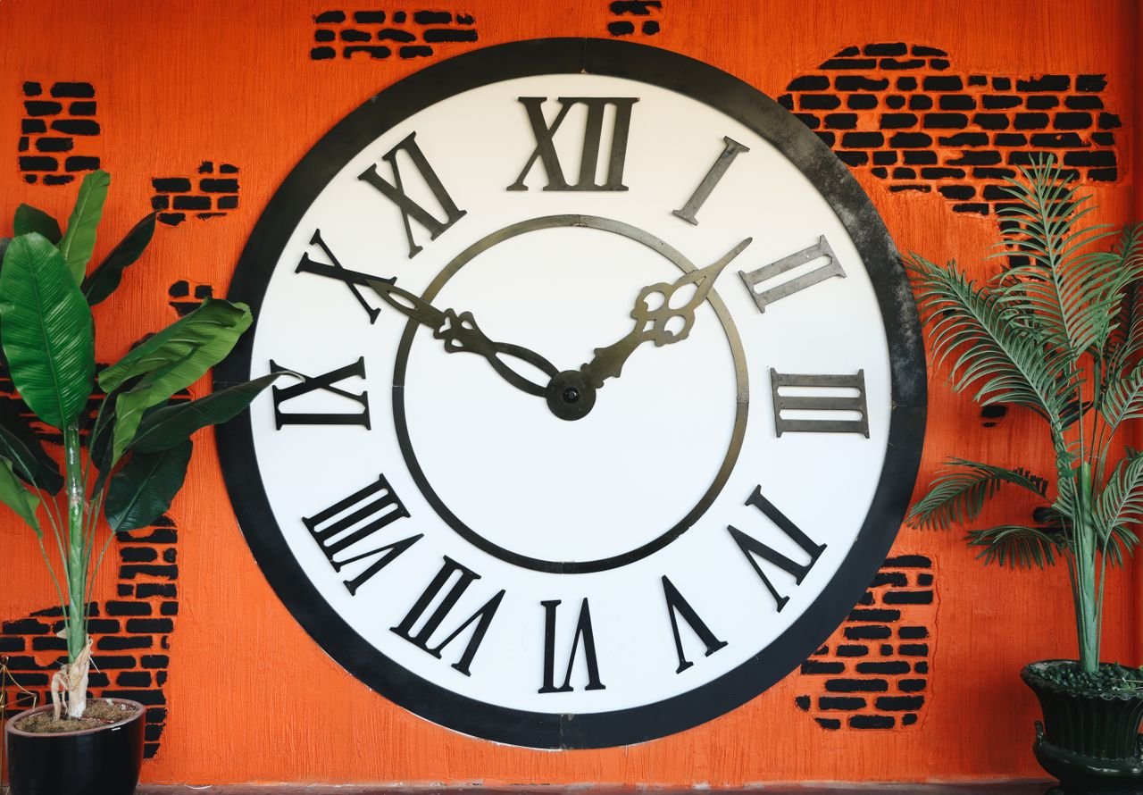A vintage-style wall clock with Roman numerals against a vibrant orange brick background.